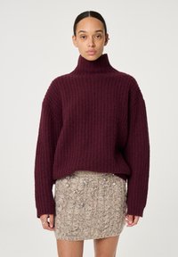 Burgunderfarbener Strickpullover mit hohem Kragen, kombiniert mit einem beigen, strukturierten Rock mit Verzierungen. Große, lässige Passform und gerippte Muster.