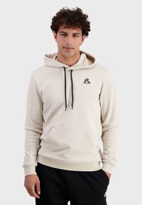 Sudadera crema de tejido suave, con un bolsillo canguro, cordón ajustable y un pequeño logo negro en el pecho.