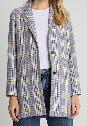 Nainen, jolla on harmaa ruudullinen bleiseri, jossa keltaiset ja mustat raidat, valkoinen paita ja siniset farkut.