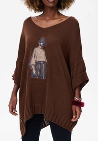 Bruine gebreide poncho met een relaxte pasvorm, met een grafisch ontwerp van een gestreepte figuur. Ribbeldetails op de mouwen en de zoom, textuurstof.