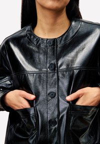 Veste en simili cuir noir avec un col rond, cinq boutons, grandes poches et une finition texturée.