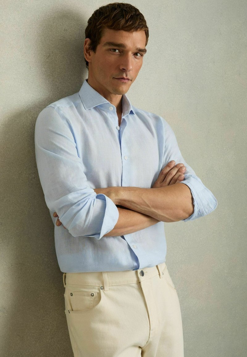 Camisa de lino azul claro con cierre de botones, puños enrollados y pantalones color crema. El conjunto presenta un diseño minimalista y texturas suaves.