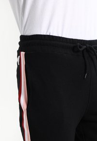 Close-up van zwarte trainingsbroek met een koord in de taille en verticale zijstrepen in rood, wit en roze, gedragen met een wit shirt.
