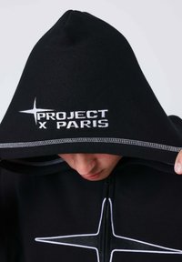 Sudadera negra con cremallera en la parte delantera, que presenta "PROJECT x PARIS" bordado en blanco en la capucha, acentuado por costuras contrastantes.