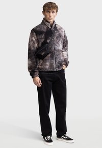 Faux-Fur-Jacke mit einem marmorierten Muster in Dunkelbraun und Grau, schwarzer Reißverschluss vorne, kombiniert mit schwarzen Hosen und einer schwarzen Umhängetasche.
