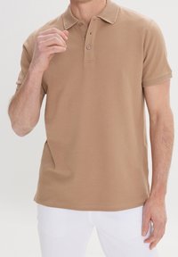 Polo shirt camel à manches courtes en coton doux avec un col texturé et une patte de boutonnage à trois boutons, associé à un pantalon blanc.