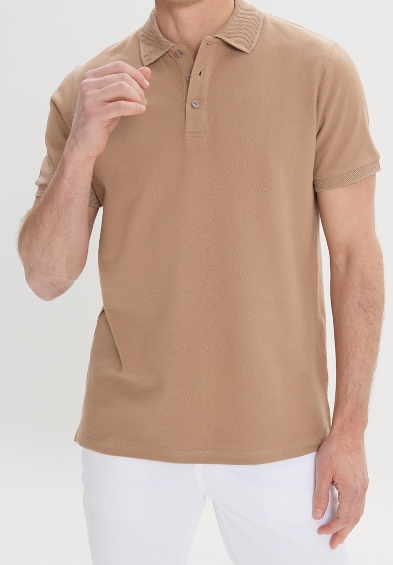 Polo shirt camel à manches courtes en coton doux avec un col texturé et une patte de boutonnage à trois boutons, associé à un pantalon blanc.