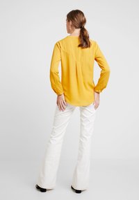 Gelbe Bluse mit langen Ärmeln und rundem Kragen, gerafften Ärmeln und Rückennaht, kombiniert mit weißen Schlaghosen und schwarzem Schuhwerk.