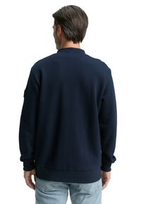 Granatowa bluza z teksturowanej tkaniny; ma okrągły dekolt oraz ściągacze na mankietach i u dołu. Noszona z jasnoniebieskimi dżinsami.