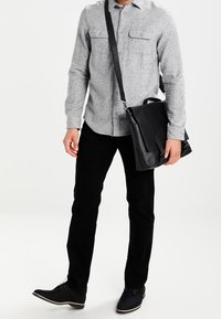 Černá kožená taška přes rameno s klopou, nošená jako crossbody. Šedá texturovaná košile se dvěma náprsními kapsami, doplněná černými kalhotami a botami.