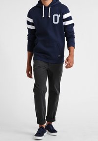 Sudadera con capucha azul marino y mangas a rayas blancas, con bolsillo frontal, combinada con pantalones oscuros ajustados gris oscuro y zapatos oscuros.