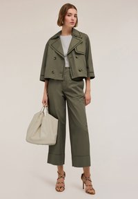 Olivengroene cropped jas met dubbele knopen, gecombineerd met bijpassende wijde broek. Model houdt een crèmekleurige handtas vast en draagt sleehakken.