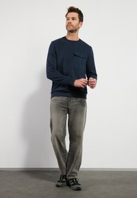 Donkerblauwe longsleeve sweatshirt met een borstzak, grijze spijkerbroek en zwarte sportschoenen. Het sweatshirt heeft een gladde textuur en een relaxte pasvorm.
