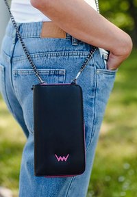 Black leather crossbody bag with a pink zipper accent, rectangular shape, and chain strap, attached to a person’s denim jeans.