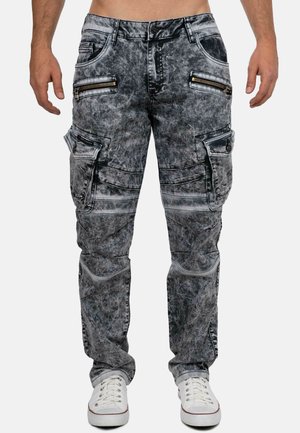 Mann trägt graue Acid-Wash-Cargohosen mit Reißverschluss- und Klappentaschen, steht mit weißen Sneakers auf einfachem Hintergrund.