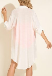 Kobieta z blond włosami ubrana w luźną, białą sukienkę z długim rękawem i podwiniętymi rękawami, stojąca na jednolitym jasnym tle.