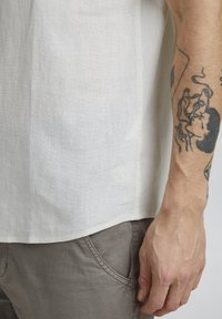 Chemise en lin crème à manches courtes avec un tissage texturé, présentant un détail de logo discret sur l'ourlet et associée à un pantalon gris clair.