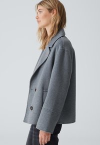 Graue Wollmischungjacke mit Reverskragen, doppelreihiger Knopfleiste und seitlichen Taschen. Weiche Textur und lässige Passform mit sichtbaren Nähten.