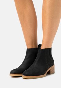 Bottines en suède noir avec panneaux latéraux élastiques, bouts ronds et un talon en bois empilé. Couture visible le long de la semelle.