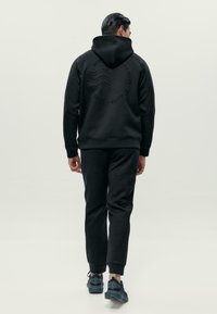Muž odchádzajúci v čiernej mikine s kapucňou a zodpovedajúcich čiernych teplákových nohaviciach so čiernymi teniskami na jednofarebnom svetlom pozadí.