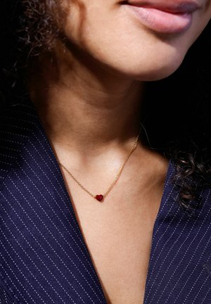 Gold chain necklace featuring a small red heart pendant, worn on a bare neck, against a striped navy fabric background.