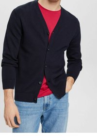 Marinblå cardigan med ribbat mönster, sex knappar framtill och långa ärmar. Bärs över en röd skjorta och matchas med ljusblå jeans.