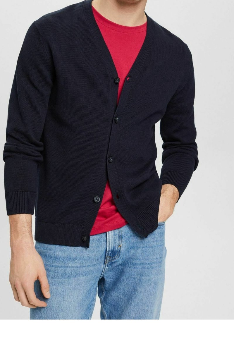 Marinblå cardigan med ribbat mönster, sex knappar framtill och långa ärmar. Bärs över en röd skjorta och matchas med ljusblå jeans.