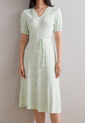 Femme portant une robe fleurie vert clair, longueur genou, à col en V, manches courtes bouffantes, et ceinture nouée à la taille, debout devant un mur uni.