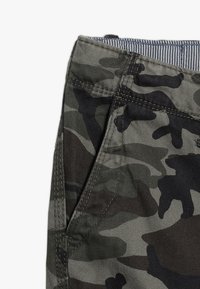 Camouflagegroen stof met een gestructureerd patroon, zichtbaar tailleband en stikseldetails. Close-up van een zaknaad.