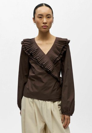 Kobieta ubrana w brązową, długorękawową bluzkę kopertową z plisowanymi falbanami oraz kremowe spodnie o szerokich nogawkach.