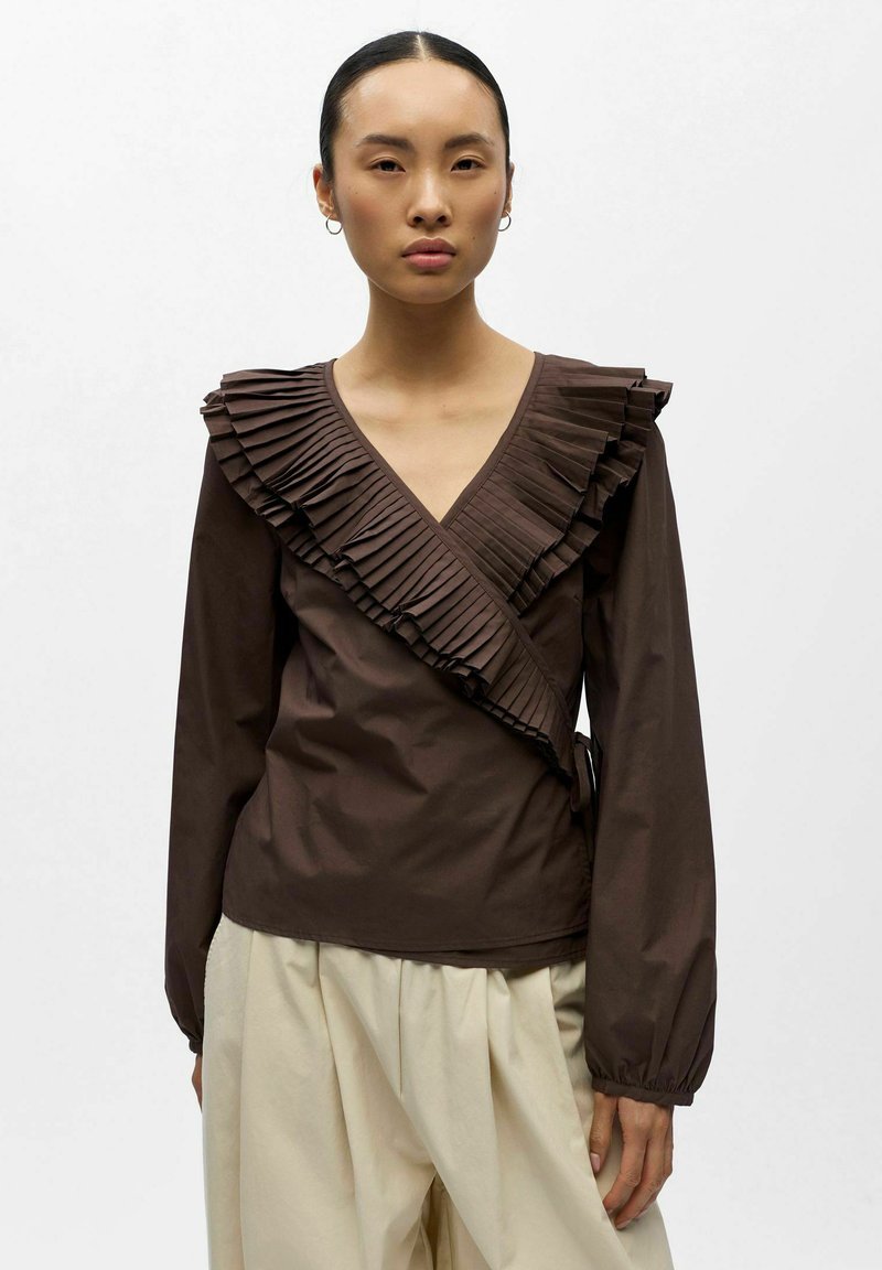 Femme portant un haut cache-cœur à manches longues marron avec des détails plissés et volantés, ainsi qu'un pantalon large de couleur crème.