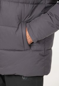 Grå vadderad jacka med en strukturerad yta, med en sidficka och en åtsittande design. Inga synliga logotyper eller mönster.