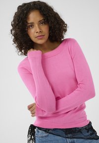 Mujer con cabello rizado vistiendo una blusa de mangas largas rosa brillante y jeans azules, posando con una mano cerca de su rostro y un fondo neutro.