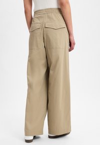 Beige wijde broek met een elastische tailleband en twee achterzakken. De stof heeft een soepele textuur en een lichte uitstraling.