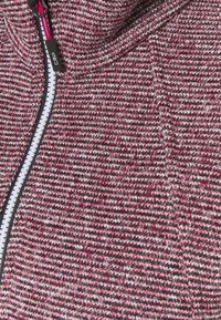 Tissu en maille texturée dans des tons de rose et de gris avec une fermeture éclair blanche proéminente. Doté d'une capuche et d'un motif discret sur l'ensemble.