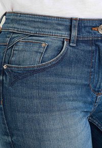 Jean en denim d'un bleu moyen avec des coutures contrastantes orange, présentant une poche avant et une taille classique.