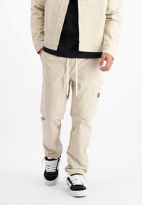 Beige cargobroeken met elastische tailleband en trekkoord, voorzien van zijzakken en een gestructureerde stof. Draag het met een zwart shirt en sneakers.