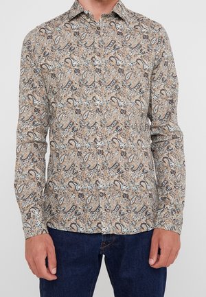 Mann trägt ein beige Paisley-Print langärmliges Hemd mit dunklen blauen Jeans, steht vor einem einfarbigen Hintergrund.