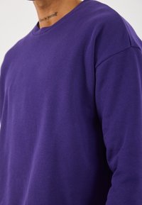 Man met een effen paarse lange mouw shirt, zichtbaar nek tatoeage met de woorden "geloof in jezelf."