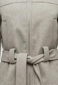 Gros plan d’un manteau en laine gris clair avec une fermeture éclair verticale et une ceinture nouée autour de la taille.