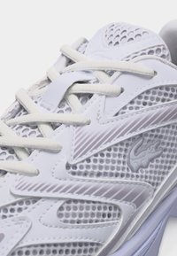 Λευκό αθλητικό παπούτσι με πάνελ από δίχτυ, επικαλυπτόμενα λευκά κορδόνια και ένα μικρό ανάγλυφο λογότυπο κροκοδείλου στο πλάι.