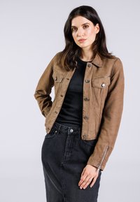 Veste en cuir suédé marron avec fermeture boutonnée, deux poches poitrine et une coupe ajustée. Associée à un haut noir à côtes et une jupe en denim foncé.
