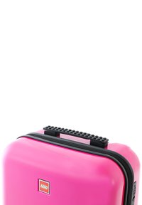 Hard-shell pink suitcase with black zipper and handle. Top features raised ridges resembling LEGO bricks. Display logo on front panel.