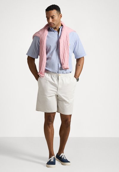 Chemise à rayures bleu clair à manches courtes, shorts crème, pullover en tricot rose enroulé autour des épaules, baskets navy avec lacets blancs.