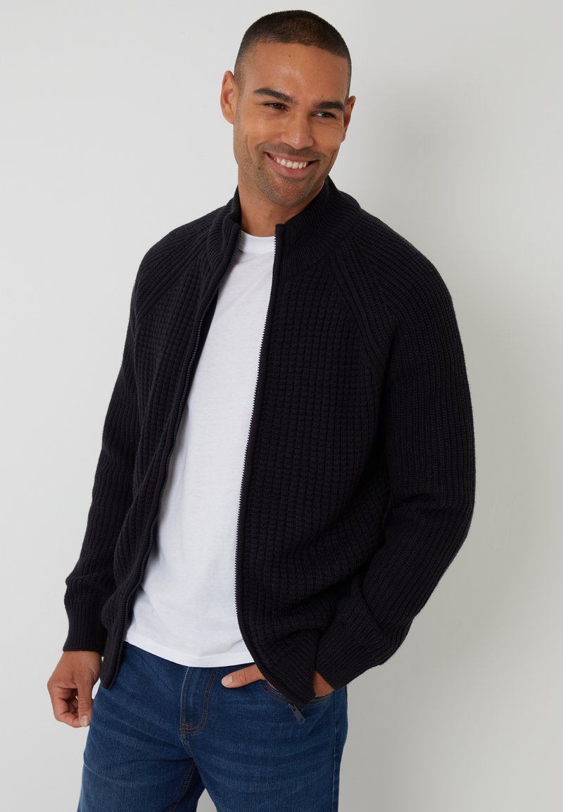 Dunkelblau gestrickte Zip-Up-Cardigan mit einem strukturierten Muster, geripptem Kragen und enganliegenden Ärmeln, getragen über einem weißen T-Shirt.