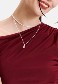 Silberne Lagenhalsketten, getragen über einem bordeauxfarbenen schulterfreien Oberteil. Die untere Halskette trägt einen kleinen Kristallanhänger. Weiche Stofftextur.