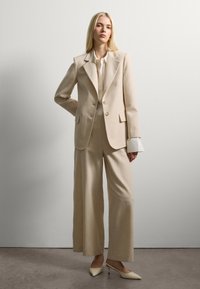Femme portant un blazer beige ajusté avec un pantalon large assorti, un chemisier crème et des escarpins à bout pointu, se tenant contre un mur uni.