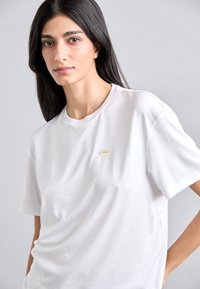 Tricou alb cu mânecă scurtă din bumbac, având un mic logo auriu în zona stângă a pieptului, cu o croială lejeră și o textură moale.