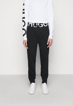 Osoba nosząca czarne spodnie dresowe z białym paskiem "HUGO", białą koszulę z długim rękawem z czarnym napisem na rękawie oraz białe trampki.