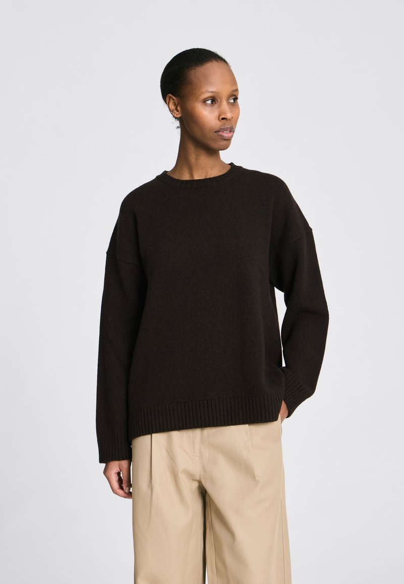 Rövid hajú nő laza fekete pulóverben és bézs színű, szélű bő nadrágban, balra néz egy egyszerű világos háttér előtt.